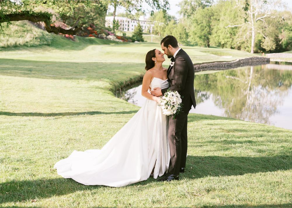 Bride in a strapless Lea-Ann Belter Arden ballgown with a full skirt and cathedral train stands with her groom on a sunlit golf course lawn beside a pond, holding a white anemone bouquet.