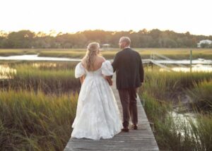 A bride and groom walk arm in arm along a wooden pier in Charleston during the golden hour. The bride wears an off the shoulder ball gown of floral organza.