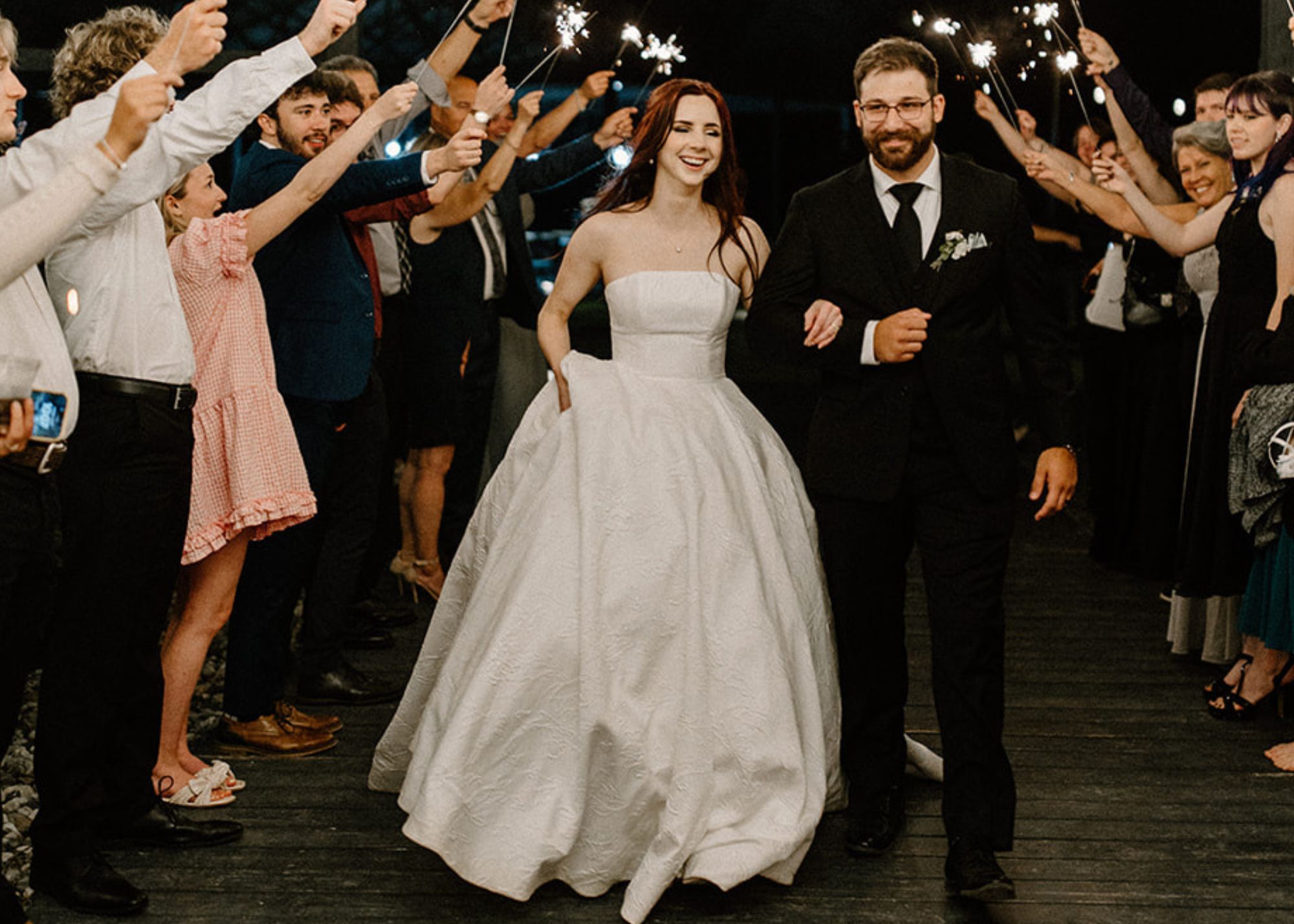 A bride and groom run arm and arm beneath sparklers. The bride wears a strapless ball gown and the groom a tux