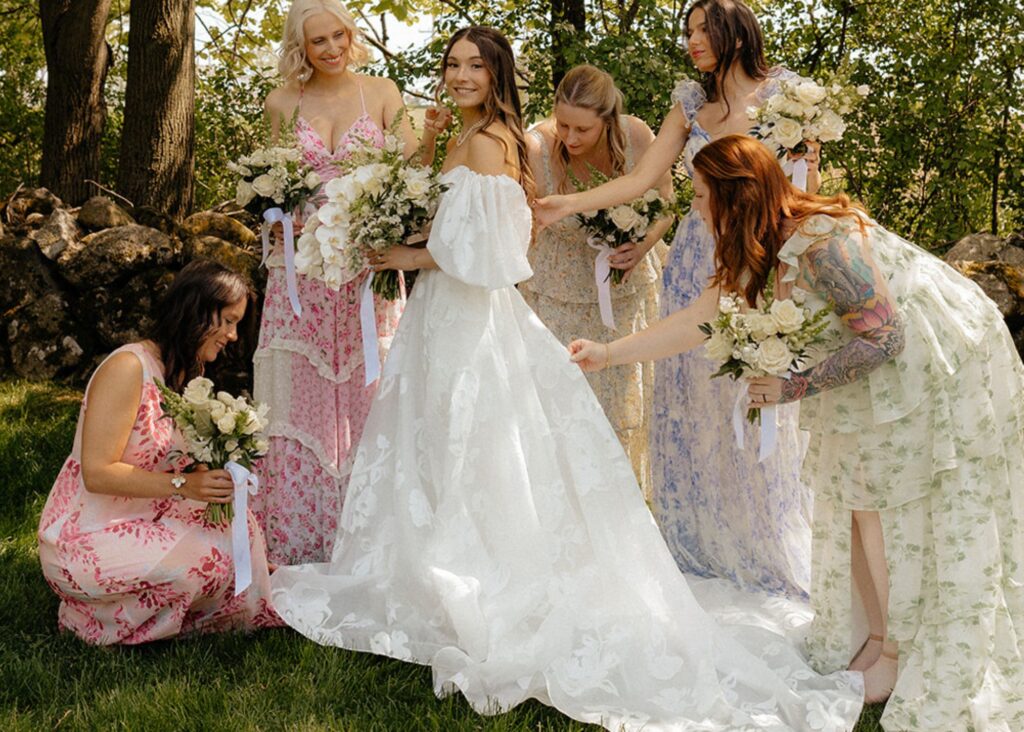 A smiling bride stands in the shade of a tree with her bridesmaids who are wearing pastel floral print dresses. The bride wears a floral organza gall gown