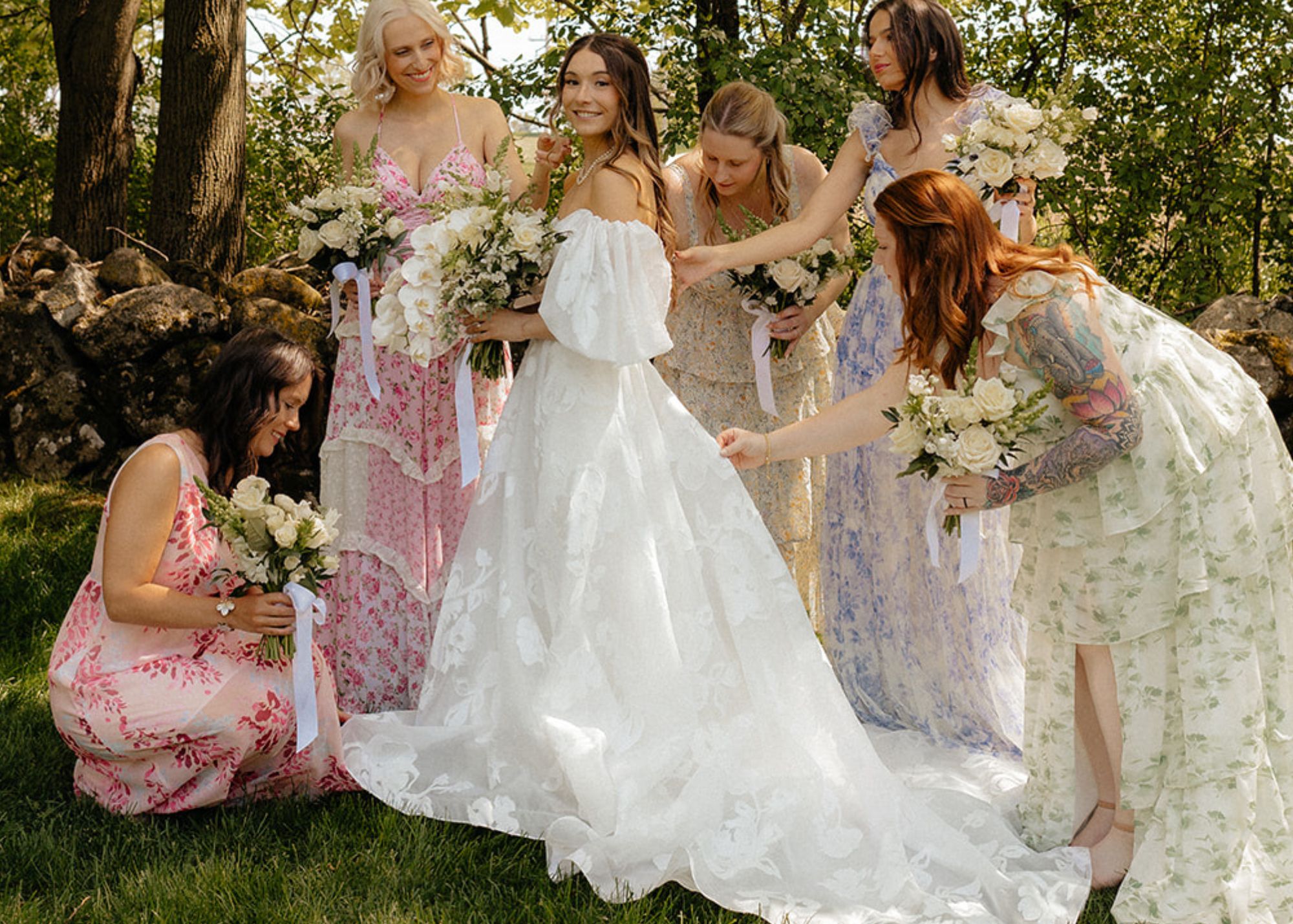 A smiling bride stands in the shade of a tree with her bridesmaids who are wearing pastel floral print dresses. The bride wears a floral organza gall gown