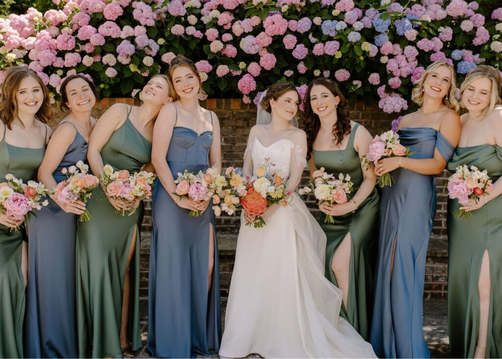 Bride in a crepe wedding gown with floral appliqué and detachable organza overskirt poses with bridesmaids in blue and green sleek gowns