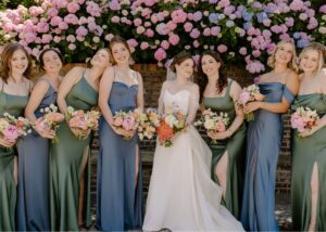 Bride in a crepe wedding gown with floral appliqué and detachable organza overskirt poses with bridesmaids in blue and green sleek gowns