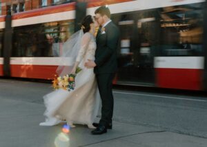 a bride and groom kiss passionately on a Toronto street as traffic rushes by. The bride wears a flowy lace wedding dress custom designed at the LABB Toronto bridal shop