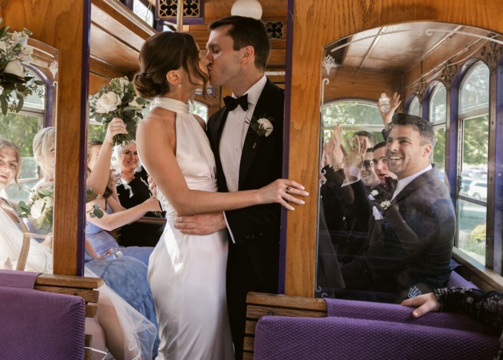 A bride and groom kiss, surrounded by their wedding party in a trolley, the bride wears a sleek wedding dress with high halter neckline by Lea-Ann Belter Bridal Toronto