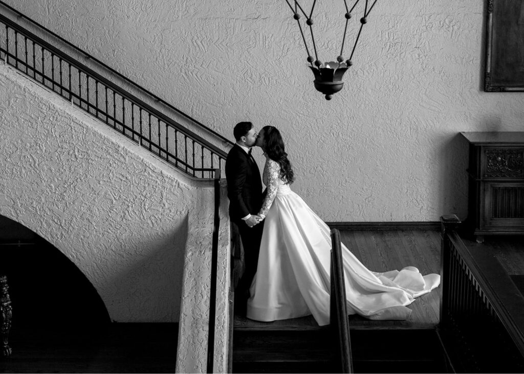 a black and white photo of a bride and groom kissing in the stairwell of Casa Loma. The bride wears a ball gown wedding dress with extra long train and tails