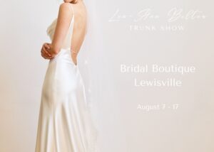 A partial shot of a bride in a low backed, bias-cut charmeuse wedding dress. Overlay text reads, Lea-Ann Belter Bridal Trunk Show at Bridal Boutique Lewisville, August 7-17