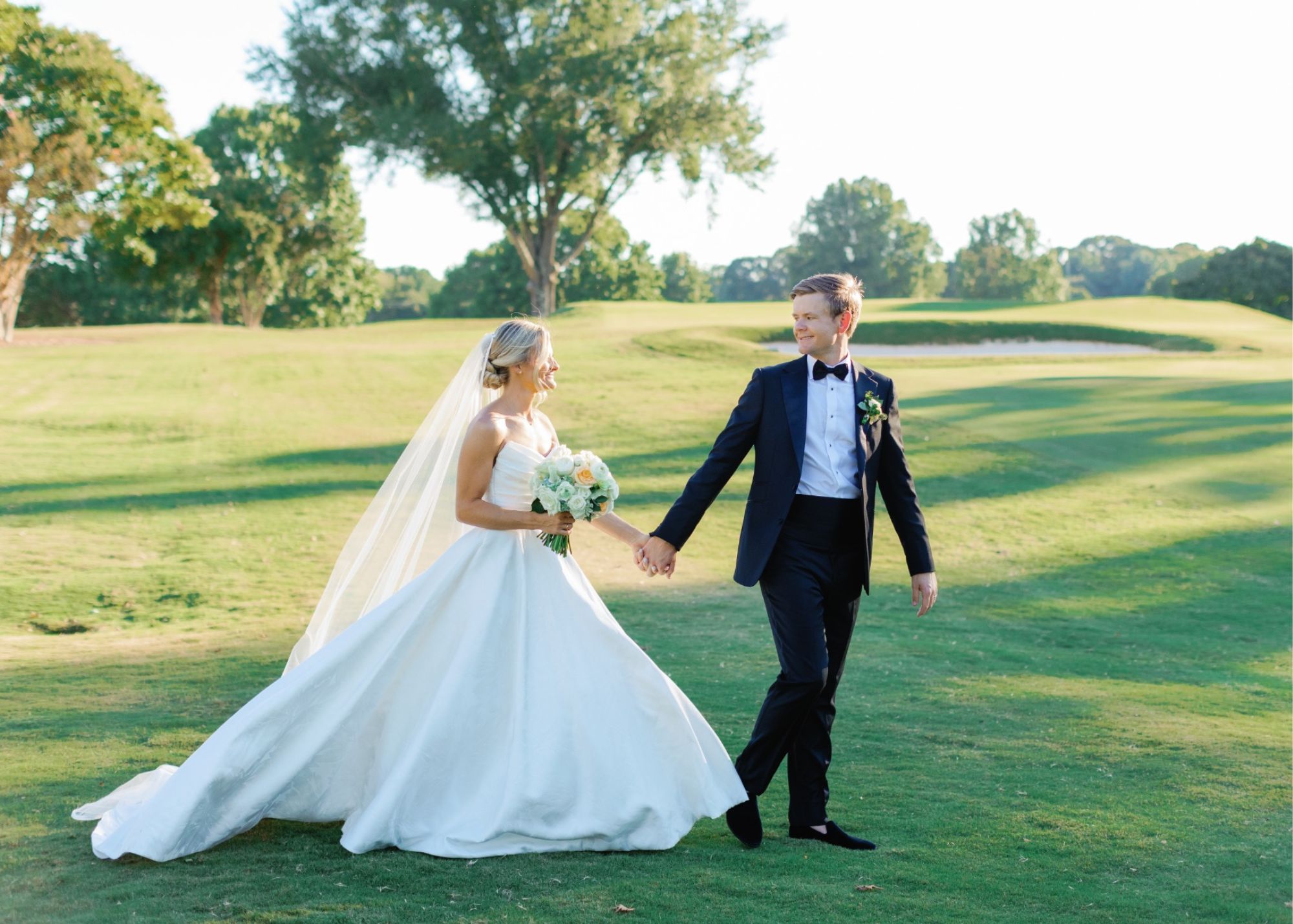 a bride in an A-line wedding dress walks with her groom in a tux across a gold course at golden hour