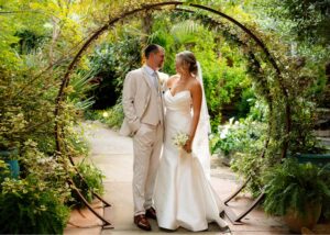 a bride and groom stand together beneath a circular arbor. He wears a tan suit, she a fit and flare wedding dress with lace edged cathedral veil