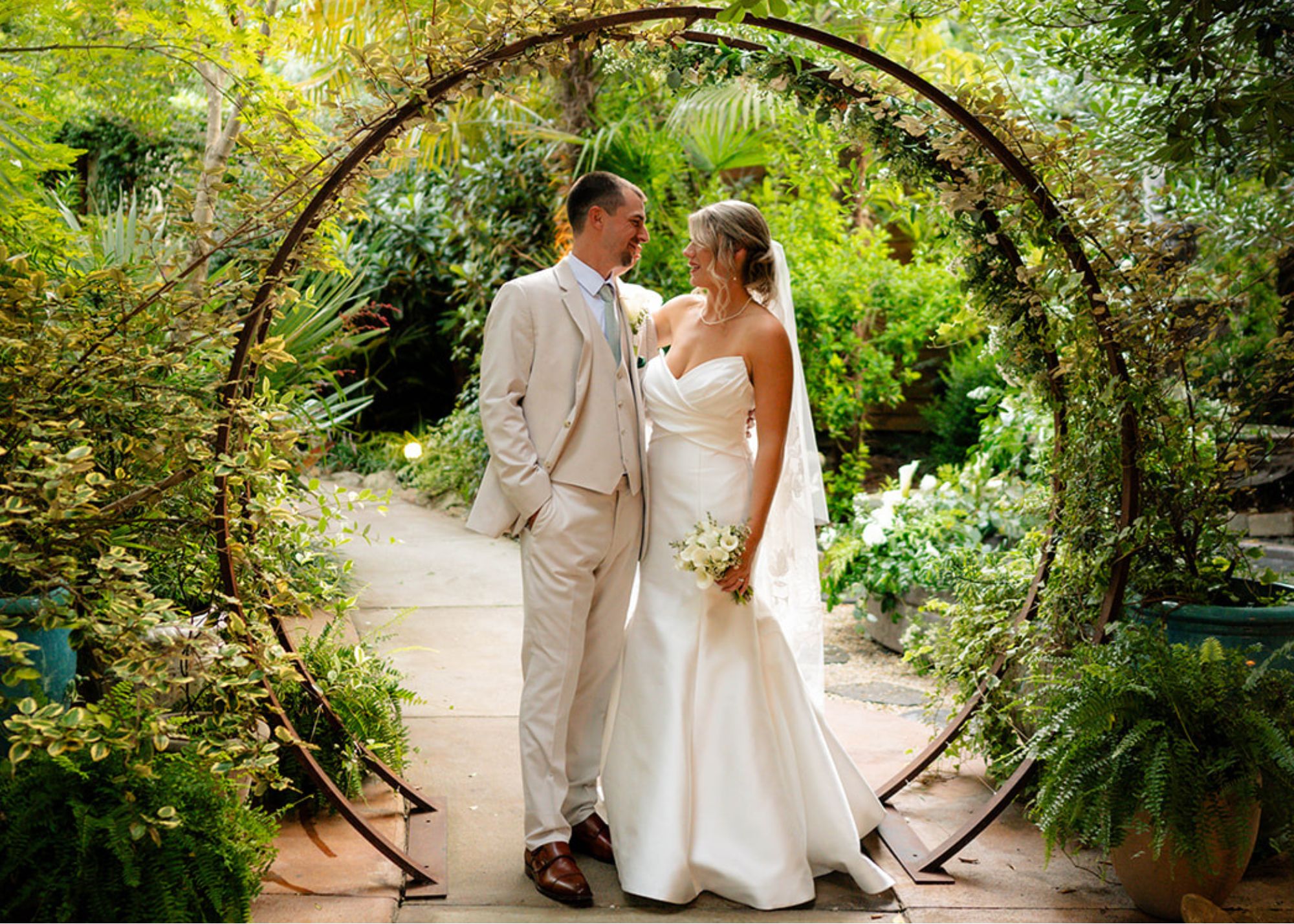 a bride and groom stand together beneath a circular arbor. He wears a tan suit, she a fit and flare wedding dress with lace edged cathedral veil