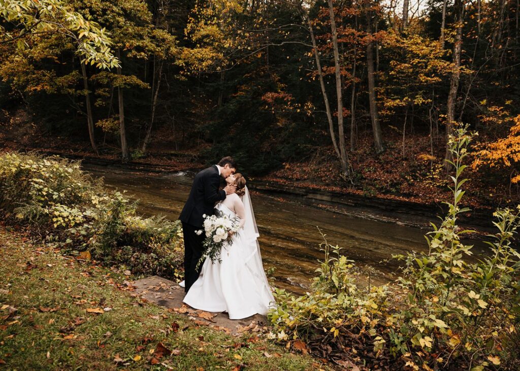 a bride and groom kiss in a garden ablaze with fall foliage. The bride wears a drop waist ball gown wedding dress with sheer net jacket with cuffs and high collar.