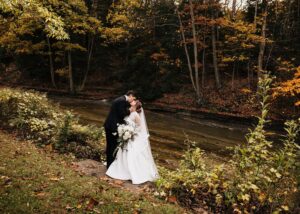 a bride and groom kiss in a garden ablaze with fall foliage. The bride wears a drop waist ball gown wedding dress with sheer net jacket with cuffs and high collar.
