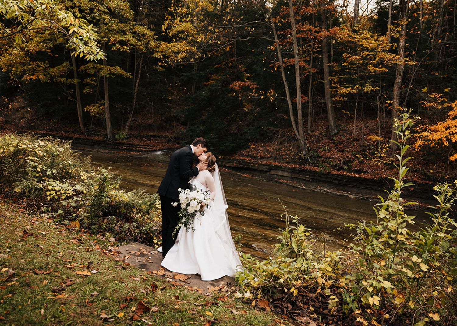 a bride and groom kiss in a garden ablaze with fall foliage. The bride wears a drop waist ball gown wedding dress with sheer net jacket with cuffs and high collar.