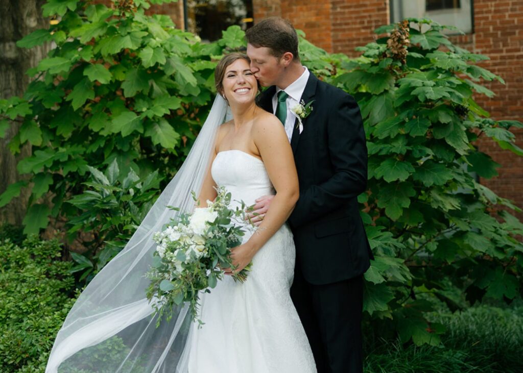 a bride and groom lean into each other, the bride wears a floral jacquard fit and flare gown and her veil flows in the breeze.