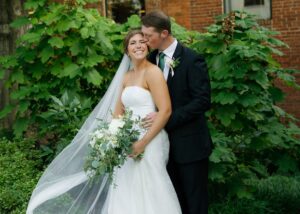 a bride and groom lean into each other, the bride wears a floral jacquard fit and flare gown and her veil flows in the breeze.