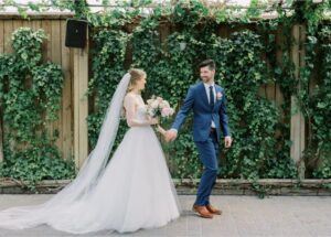 Bride in Lea-Ann Belter two piece ball gown wedding dress with cathedral veil holding hands with groom in navy suit against ivy-covered wooden fence