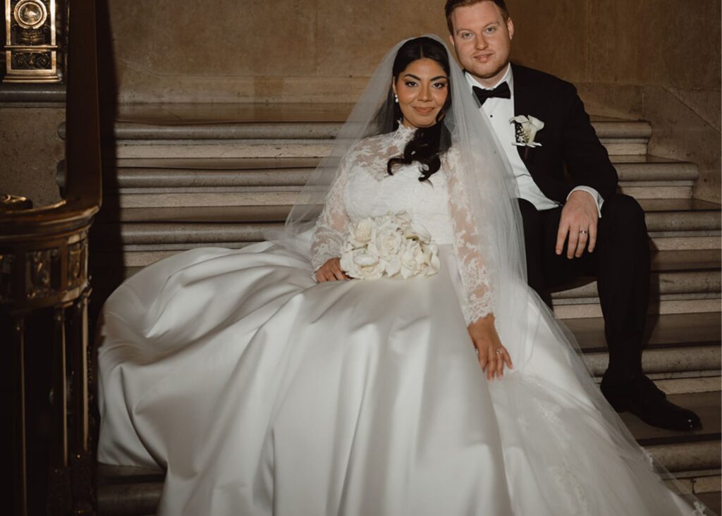 a bride in a long sleeved ball gown wedding dress and lace-edged cathedral veil sits beside her groom in a tux on a grand white marble staircase