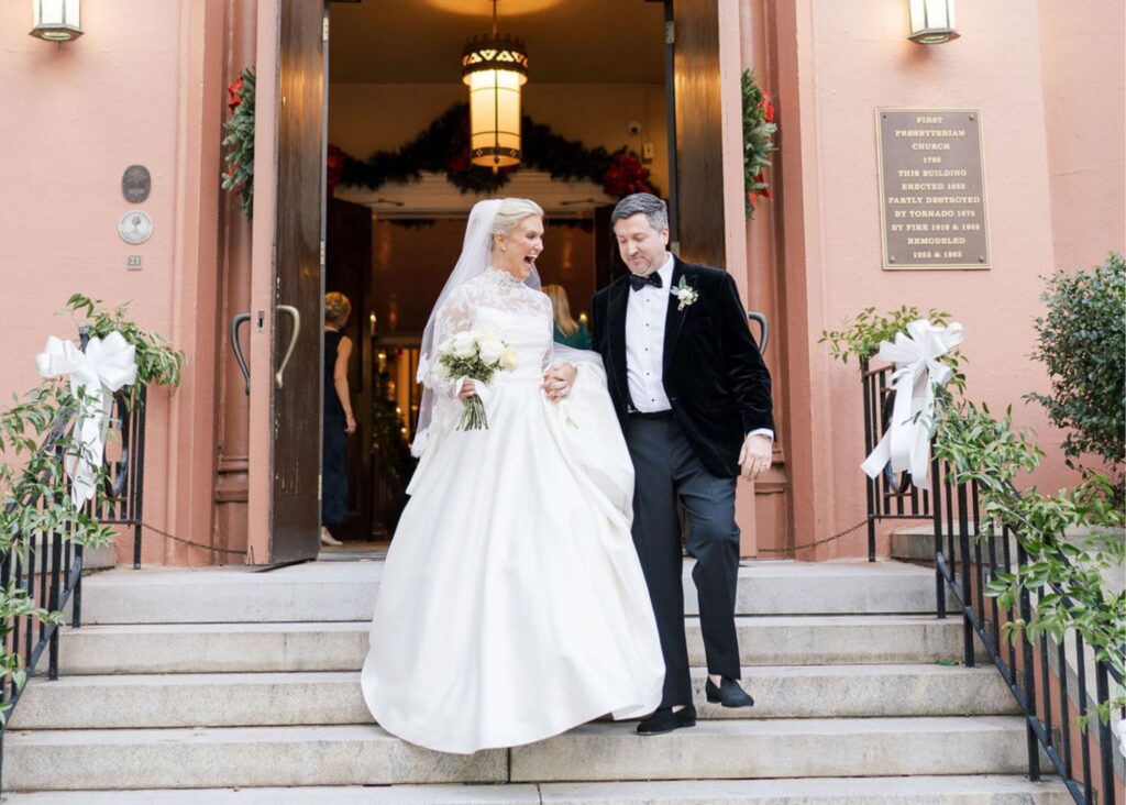 a just married bride and groom walk down the steps in front of a pink church decorated for Christmas. The bride wears a ball gown with long lace sleeves, the groom a tux