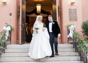 a just married bride and groom walk down the steps in front of a pink church decorated for Christmas. The bride wears a ball gown with long lace sleeves, the groom a tux