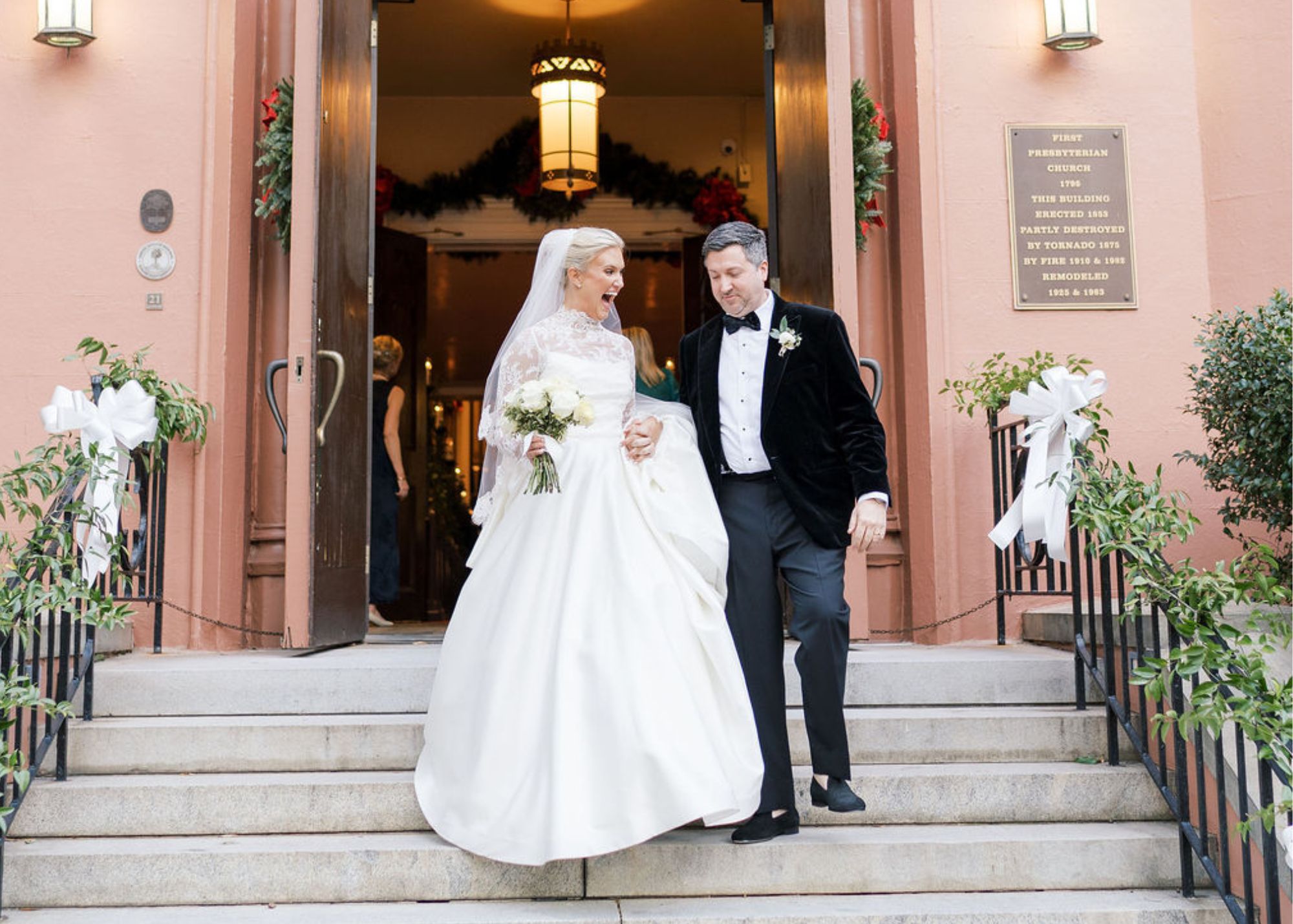 a just married bride and groom walk down the steps in front of a pink church decorated for Christmas. The bride wears a ball gown with long lace sleeves, the groom a tux