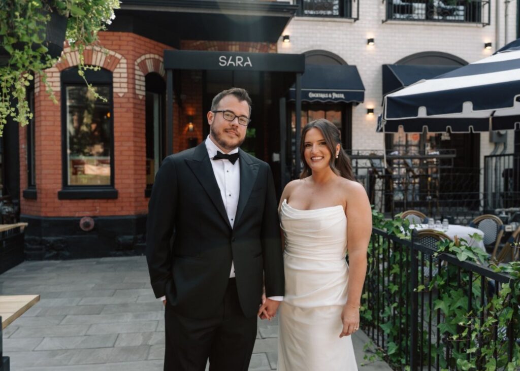 a smiling bride and groom stand in front of Toronto's Restaurant Sara. She wears a sleek wedding dress with a draped bodice, he a black tux.