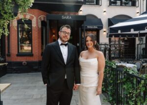 a smiling bride and groom stand in front of Toronto's Restaurant Sara. She wears a sleek wedding dress with a draped bodice, he a black tux.
