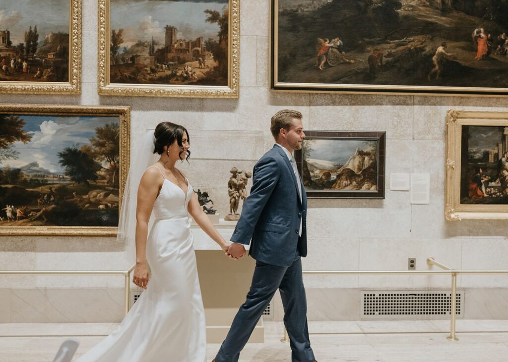 a bride and groom walk hand in hand past golf-framed paintings. She is wearing a sleek wedding dress and he a suit.