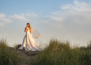 A bride stands at the top of a dune in the golden evening light, wearing an off-white lace and eco-mikado ball gown and lace-edged fingertip length veil by Toronto wedding dress designer Lea-Ann Belter