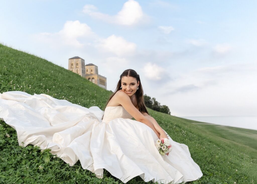 A bride sits on a grassy hill smiling, the skirts of her blush ball gown wedding dress spread about her.