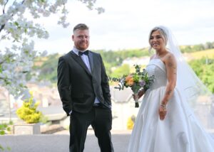 A bride and groom stand smiling at the camera at their wedding in Ireland, he in a classic suit, she in a custom ball gown by Lea-Ann Belter Toronto