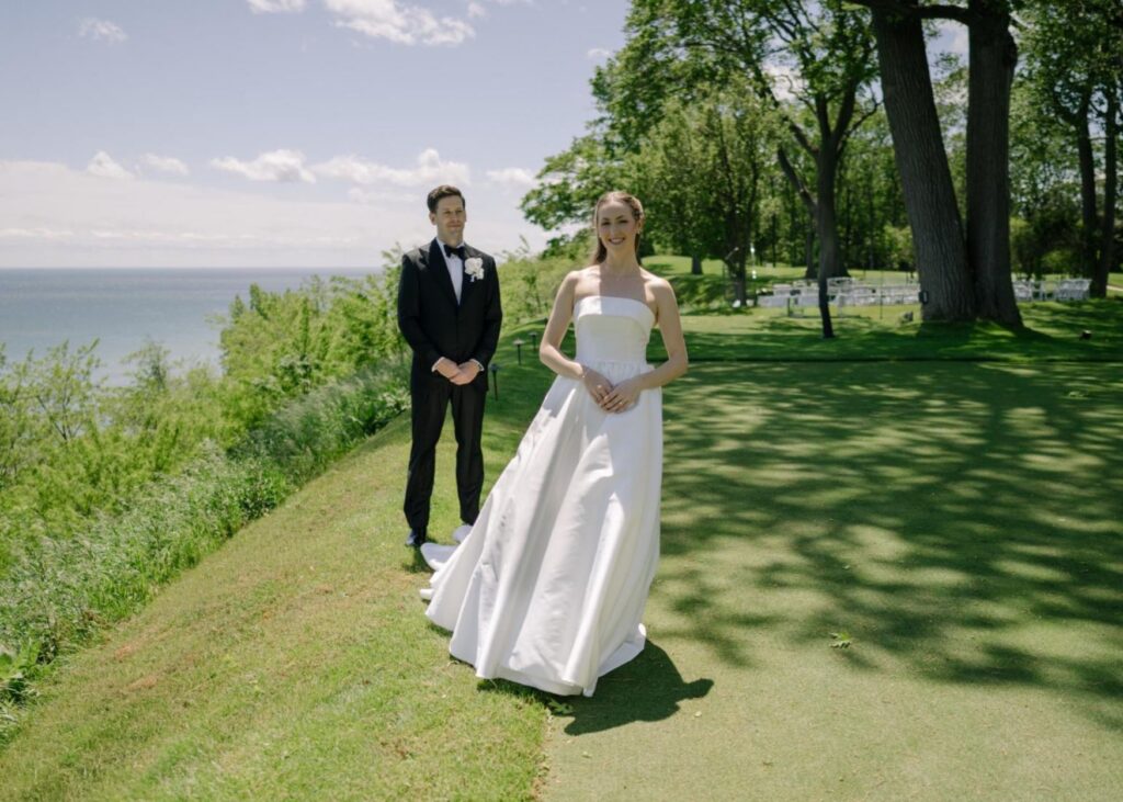 A bride and groom stand by a lake in the sunshine, a breeze flutters the skirts of the bride's bal gown wedding dress