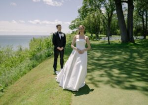 A bride and groom stand by a lake in the sunshine, a breeze flutters the skirts of the bride's bal gown wedding dress