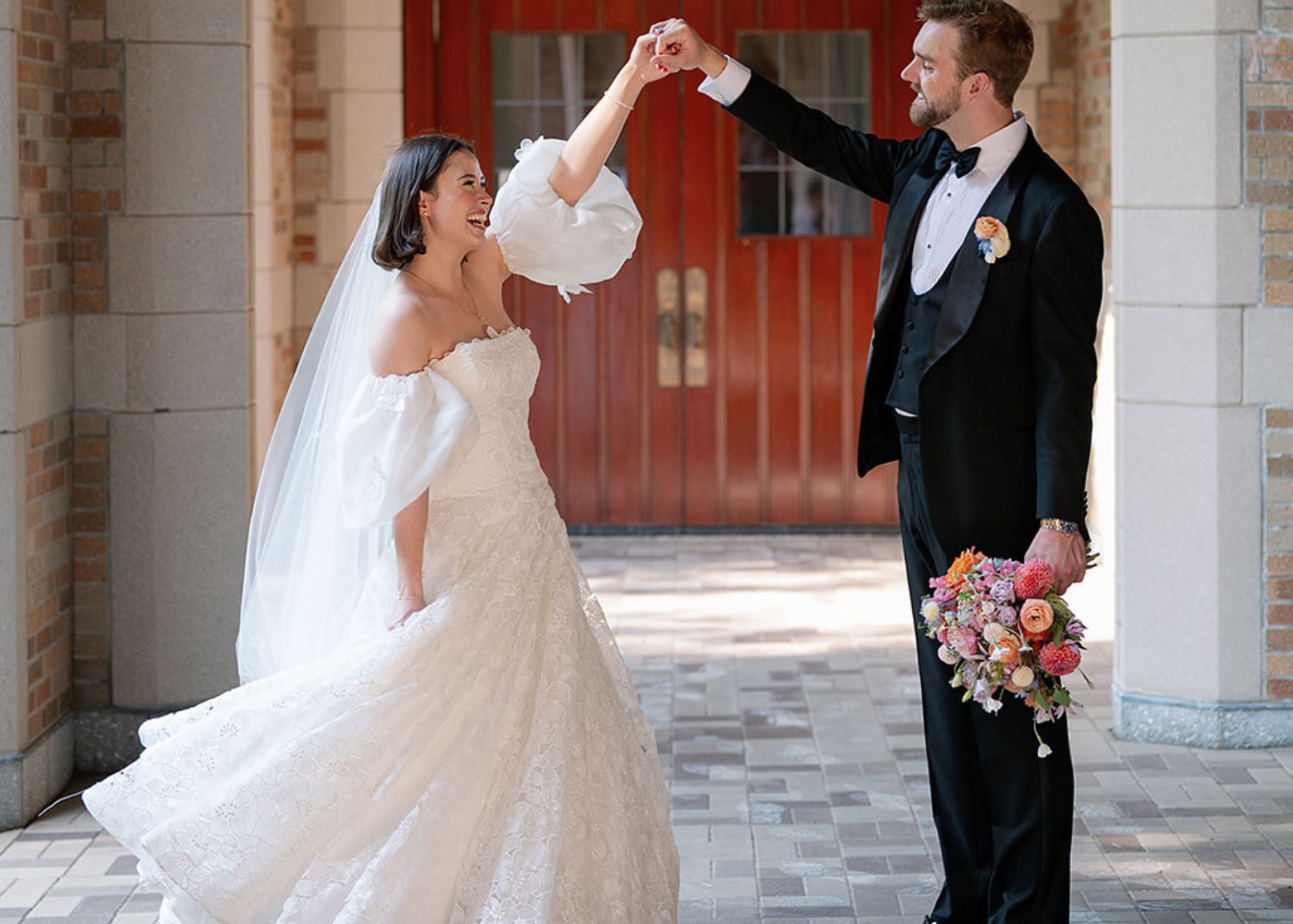 a bride pirouettes as her groom holds her hand. The bride wears a laser cut floral drop waist ball gown wedding dress with detachable puff sleeves by Lea-Ann Belter, the groom a tux.