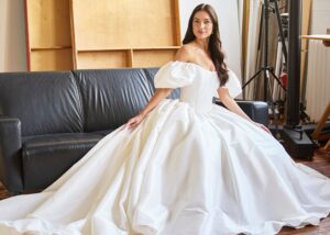 A bride with long dark hair sits ion a black couch in a ball gown wedding dress with puffed off-the-shoulder sleeves