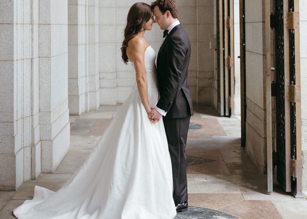 A bride and groom hold hands, foreheads together amidst columns of a St. Louis library. The groom wears a tux, the bride her hair down and drop waist ball gown of floral Jacquard by Lea-Ann Belter Bridal Toronto