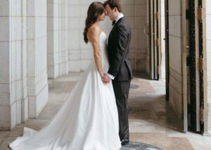 A bride and groom hold hands, foreheads together amidst columns of a St. Louis library. The groom wears a tux, the bride her hair down and drop waist ball gown of floral Jacquard by Lea-Ann Belter Bridal Toronto