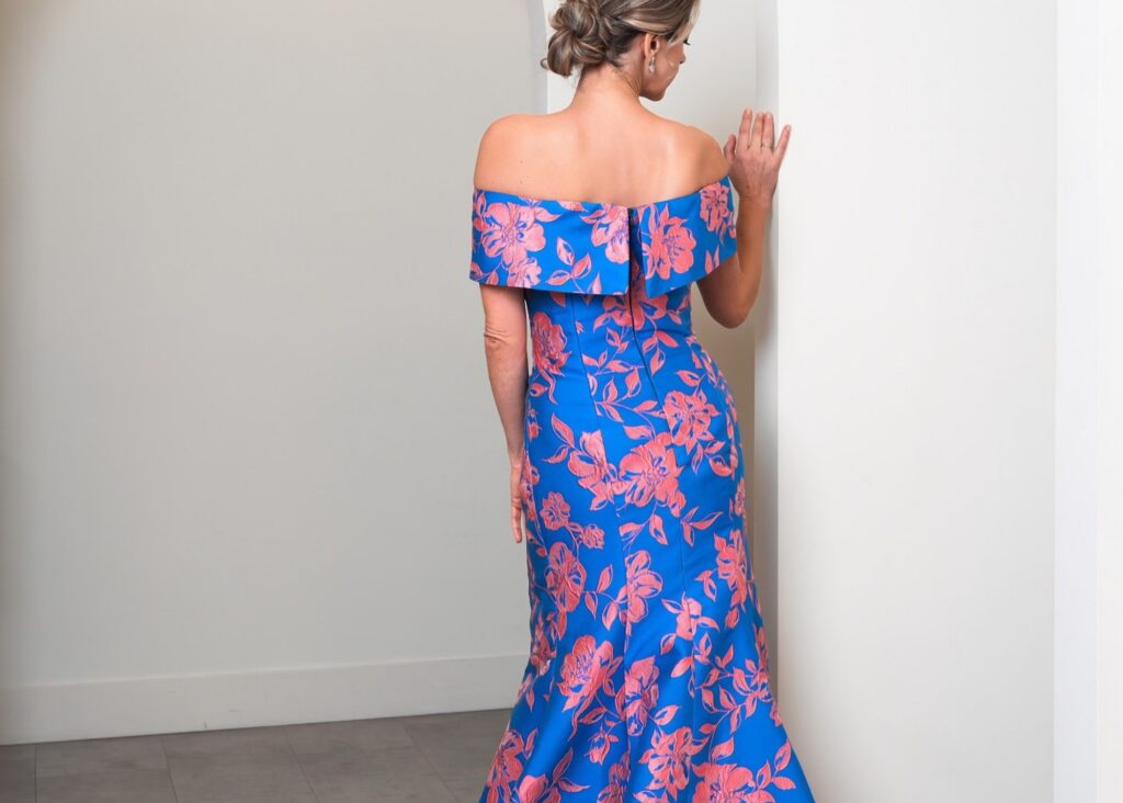 A model stands on a step with her back facing the camera wearing a a coral and sapphire floral print formal mother of the bride gown in a fit and flare shape with detachable wrap