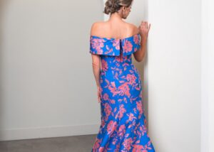A model stands on a step with her back facing the camera wearing a a coral and sapphire floral print formal mother of the bride gown in a fit and flare shape with detachable wrap