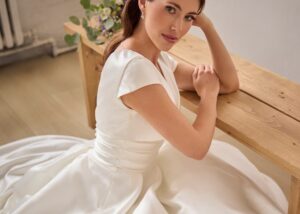 A bride wearing Peyton, a cape sleeved ball gown wedding dress, sitting on the floor in a sun-drenched studio, looking at the camera